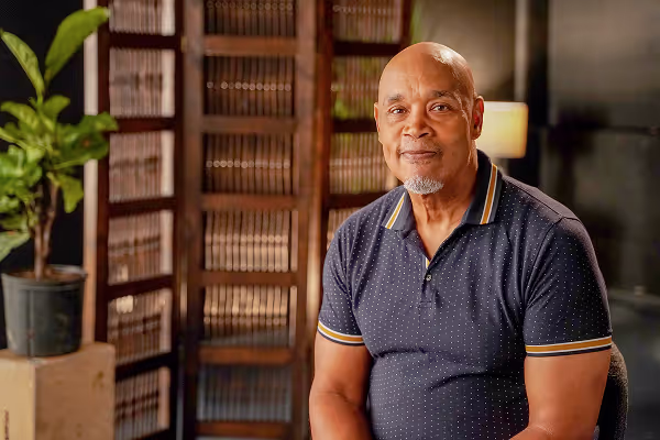 middle age man in navy blue polo sitting and smiling in a library with a plant in the background