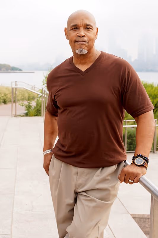 man in brown short sleeve shirt standing in an outdoor pathway with a body of water in the background   
