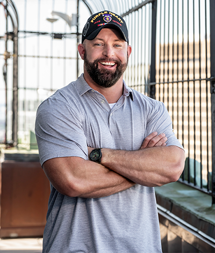 Veteran man in gray short sleeve shirt and purple heart hat with arms crossed and smiling in an office setting   