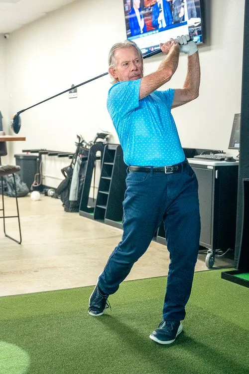 senior man in blue shirt and pants swinging golf club in an indoor golf simulator