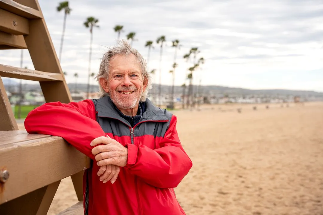senior man smiling with his arm resting on a life guard stand in a windy California beach