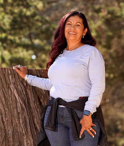 woman with dark hair and light blue shirt posing with her hand on tree trunk in the woods